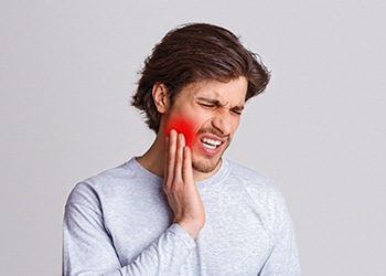 A young male wearing a gray shirt and holding his cheek in pain due to a dental injury