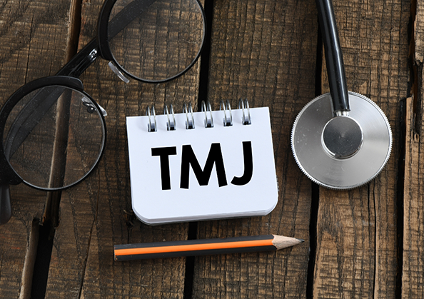Notepad that says TMJ on a desk next to a stethoscope, glasses, and a pencil